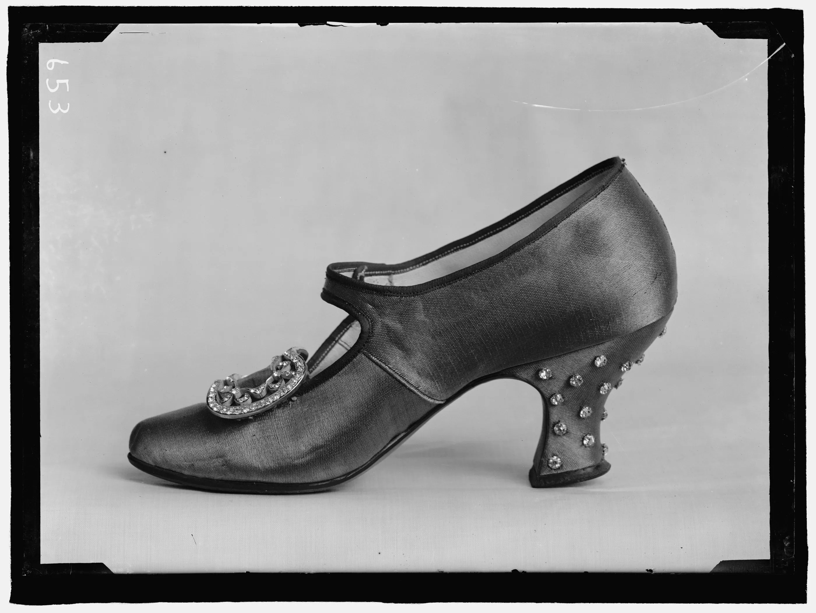 Side view of a shoe with a strap across the instep, a jewelled crescent-shaped buckle, and rhinestones set into a curved heel, photographed against a plain grey backdrop.